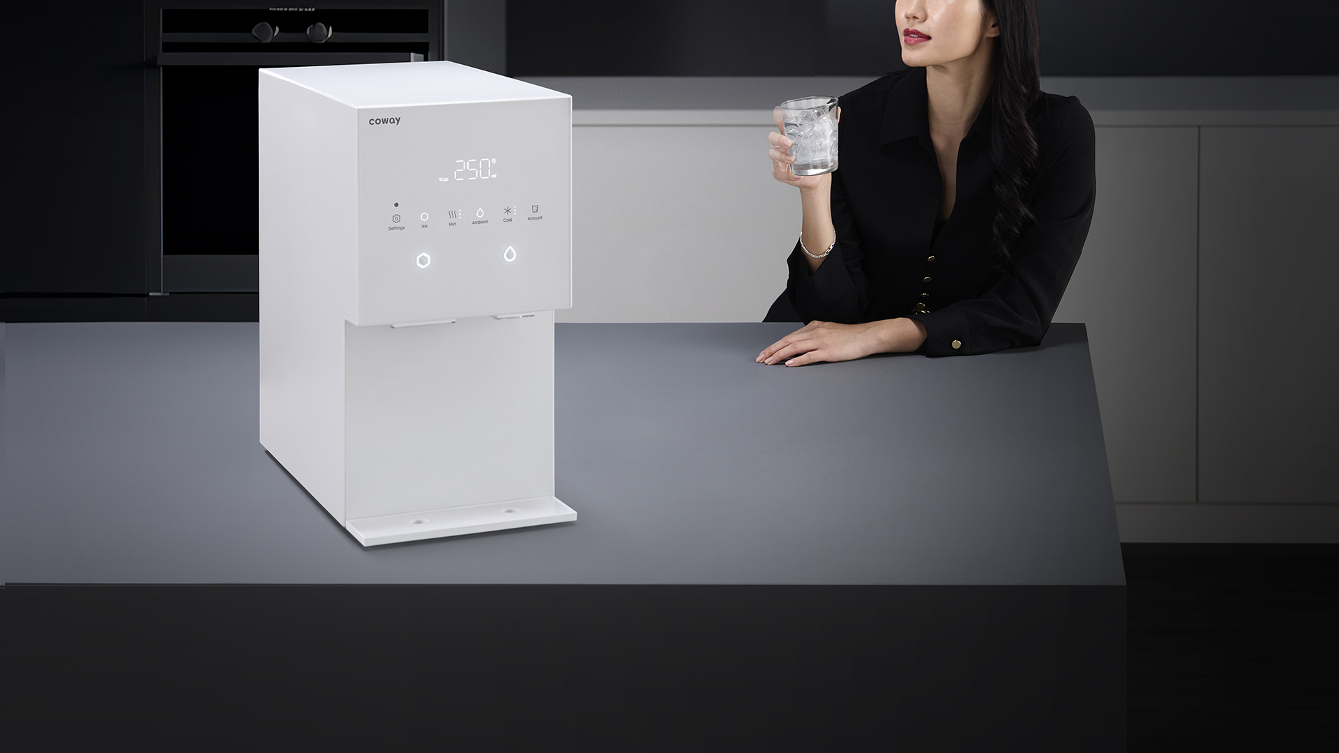 A woman enjoying a glass of iced water beside the Coway Ais in a dark modern kitchen