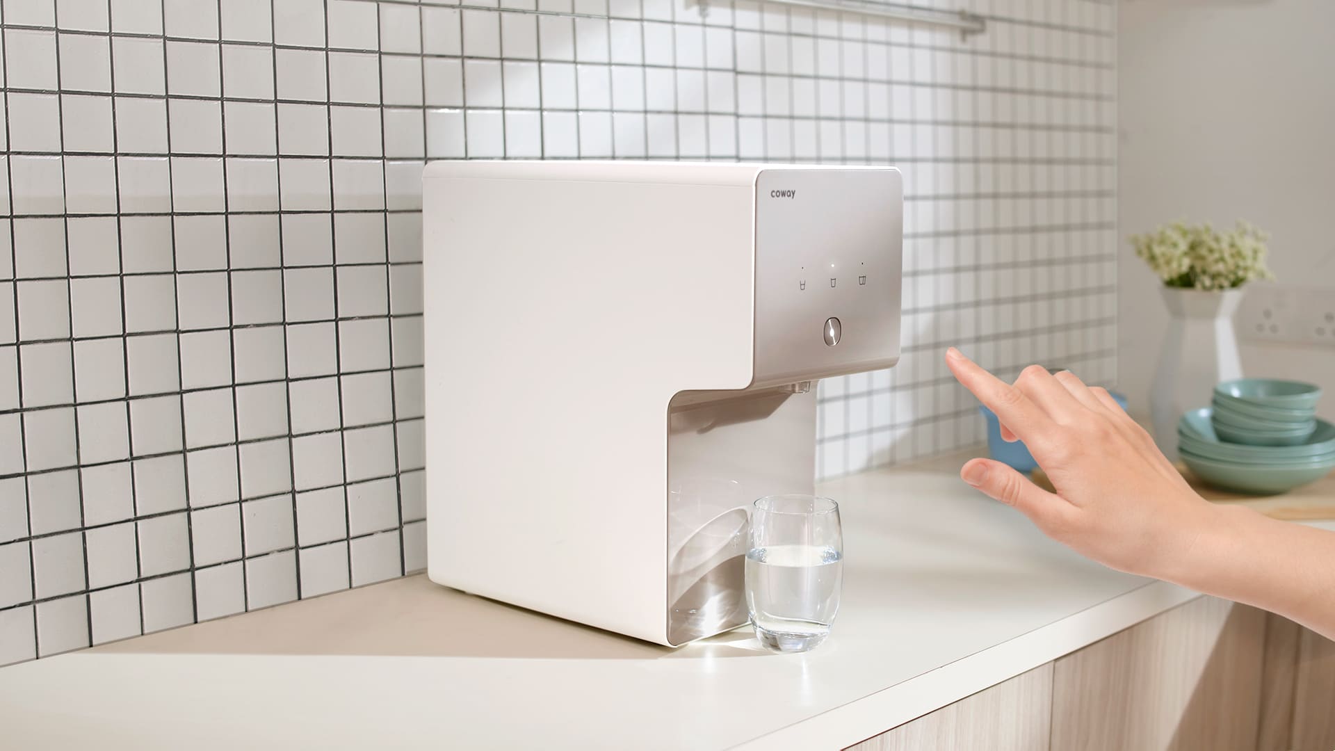 A hand reaching to touch the Coway Cinnamon dispenser in a bright tiled kitchen