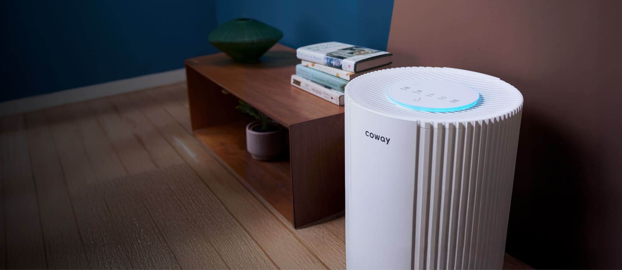 Coway Studio air purifier on a wooden floor beside a low timber console in a calm living room