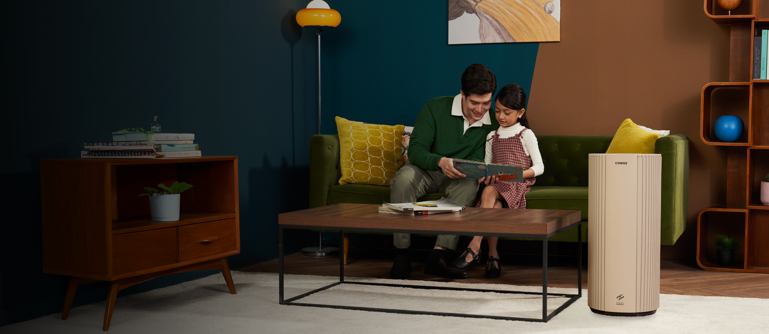 A father and daughter reading together on a green sofa with the Coway Suite air purifier beside them