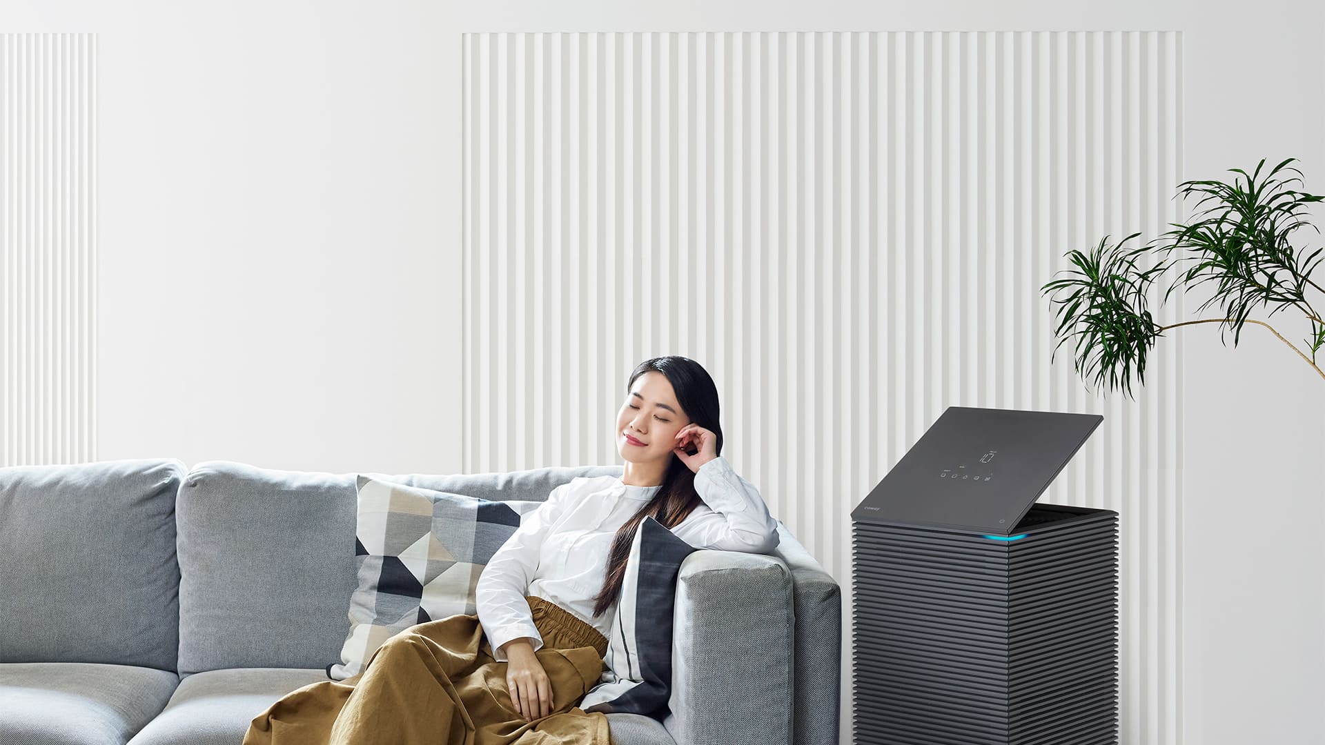 A woman relaxing beside the award-winning Coway Noble air purifier in a bright room