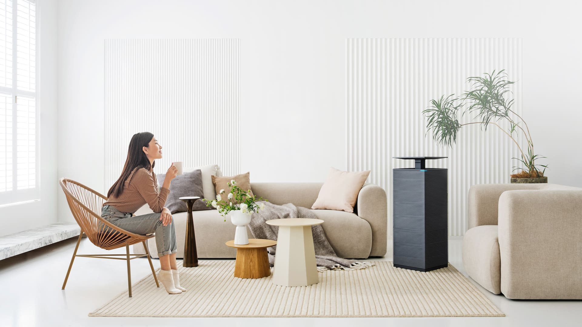 A woman relaxing with a warm drink in a bright living room beside the Coway Noble air purifier